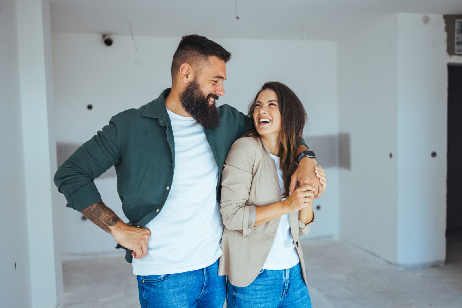 A cheerful Caucasian couple celebrates their first home purchase, standing in an unfurnished apartment with visible excitement and casual attire.
